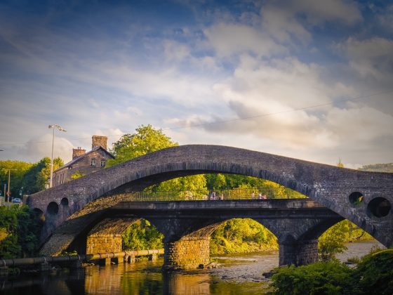 A view of Pontypridd Old Bridge: an 18th century arched stone bridge over the river Taff. Trees and houses are visible in the background, illuminated by golden evening light.