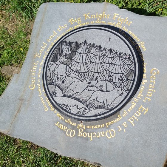 A Mab Trail story stone. The stone is set into the grass of a hillside, with a raised circular design in the centre, depicting a man on horseback hunting deer in a forest. Around the outside of the design, in gold lettering, is the title "Geraint, Enid and the Big Knight Fight". The title is repeated in Welsh.