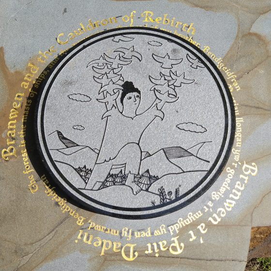 A Mab Trail story stone. The stone is set into the grass of a park, with a raised circular design in the centre, depicting a woman on a hilltop, her arms raised, dissolving into a flock of birds. Around the outside of the design, in gold lettering, is the title "Branwen and the Cauldron of Rebirth." The title is repeated in Welsh.