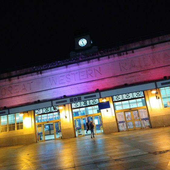 The exterior of Cardiff Central railway station at night. The building is lit in pink and yellow light. The words Great Western Railway are carved over the doors.