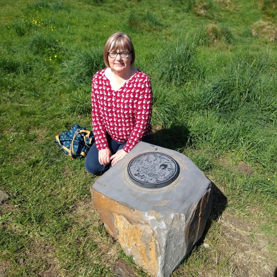 Claire Fayers, a woman with glasses and short grey/copper hair, kneels in the grass beside the stone depicting her story, "The Strange and Spectacular Dream of Rhonabwy the Restless."