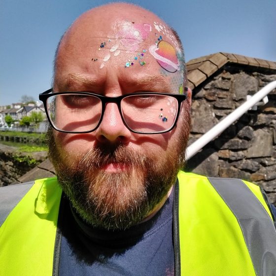 A close up photo of P.G. Bell, a middle aged man with a beard, glasses and a bald head. His face and forehead have been decorated with facepaint depicting stars and planets in bright colours. He is wearing a hi-vis vest and stands outside Pontypridd Museum in bright sunshine.