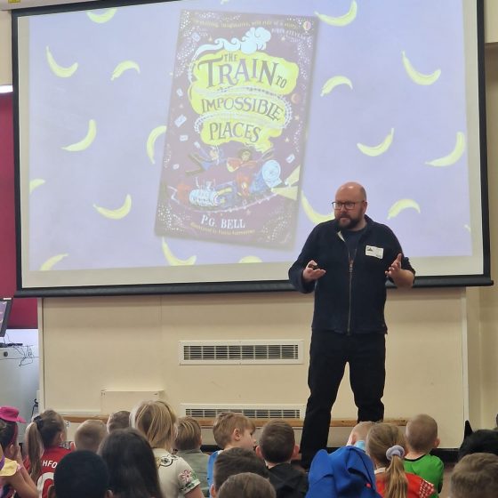 P.G. Bell talking to an audience of school children. The children are seated on the floor in the school hall, and are wearing fancy dress. The cover of The Train to Impossible Places is visible on a screen behind P.G. Bell.