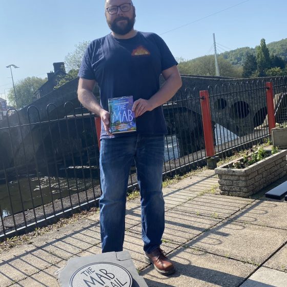 P.G. Bell, a bald, middle aged (but thoroughly good looking) man with a beard, poses with a copy of The Mab. He stands behind a story stone inscribed with "The Mab Trail". It's a bright sunny day, and the old bridge across the river Taff is visible behind him.