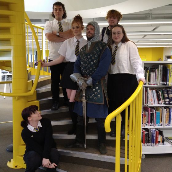 The main cast of In Time of Need pose together on a spiral staircase in the college library. They are all teenagers in school uniform, with the exception of one man dressed as a medieval knight.