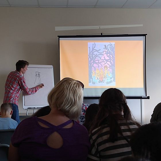 Max Low, a slim young man in a red and blue checked shirt, draws one of his Mab characters on a flipchart. An illustration from The Mab is projected onto a screen beside him. An audience sits in the foreground. We see the backs of their heads.