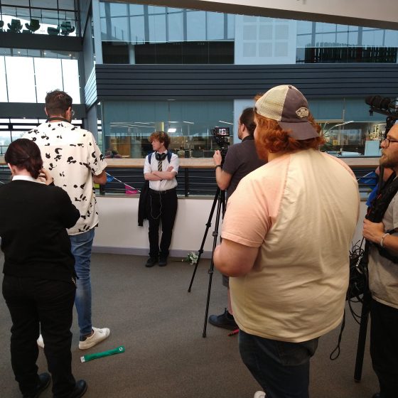 A group of crew, including a camera operator and sound man, stand on a balcony overlooking the central atrium of the college building. A young actor, Noah Jones, leans against the railings with his arms folded. He is wearing school uniform and headphones.