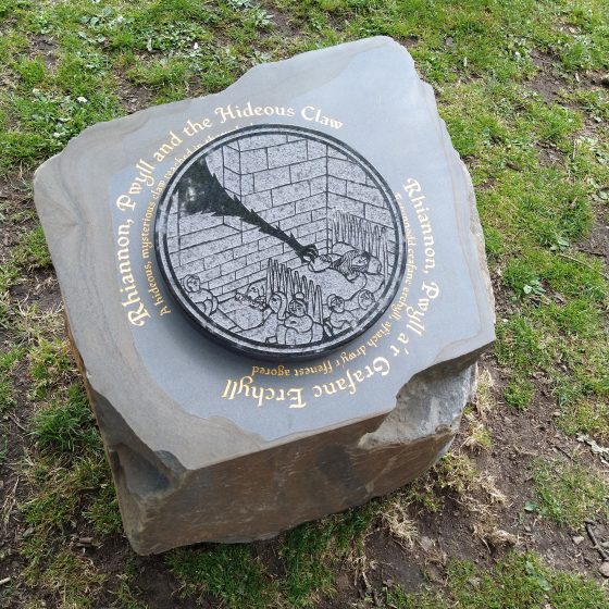 A Mab Trail story stone. The stone is set into the grass of a park, with a raised circular design in the centre, depicting a long black arm reaching through a castle window to snatch a sleeping baby. Around the outside of the design, in gold lettering, is the title "Rhiannon, Pwyll and the Hideous Claw." The title is repeated in Welsh.