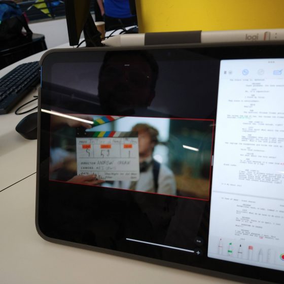 A close up of a digital tablet propped on a table. On the tablet's screen we see a feed from the camera, showing a film slate with actor Noah Jones out-of-focus in the background. To the right of the camera feed is a window displaying the script for the scene about to be filmed.