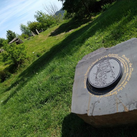 A Mab Trail story stone. The stone is set on a grassy hilltop, with a raised circular design in the centre, depicting a knight confronting a giant snake coiled around a tower of stones. Around the outside of the design, in gold lettering, is the title "Peredur, the Monster and the Serpent of the Cairn". The title is repeated in Welsh. Trees and a ring of standing stones are visible beyond the stone.