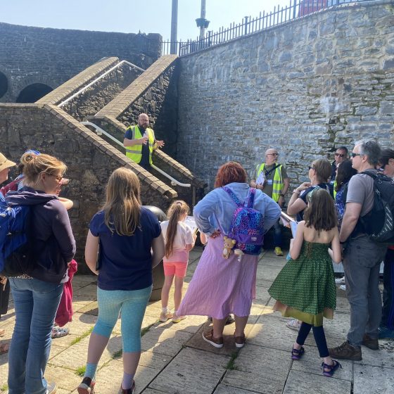 P.G. Bell, wearing a high vis vest, stands part way up a flight of stone stairs, addressing a small crowd of adults and children. They are all surrounded by high stone walls leading up towards the Old Bridge in the background.