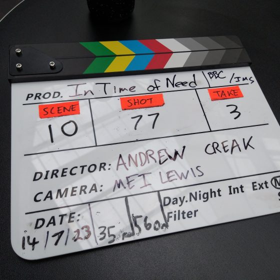 A slate or clapper board for In Time of Need. It lists Andrew Creak as the director and Mei Lewis as the camera operator. The scene marked on the slate is Scene 10, Shot 77, Take 3.
