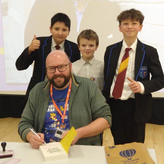 P.G. Bell sitting at a desk, signing a copy of The Train to Impossible Places. Three male pupils stand behind him, smiling and giving thumbs up