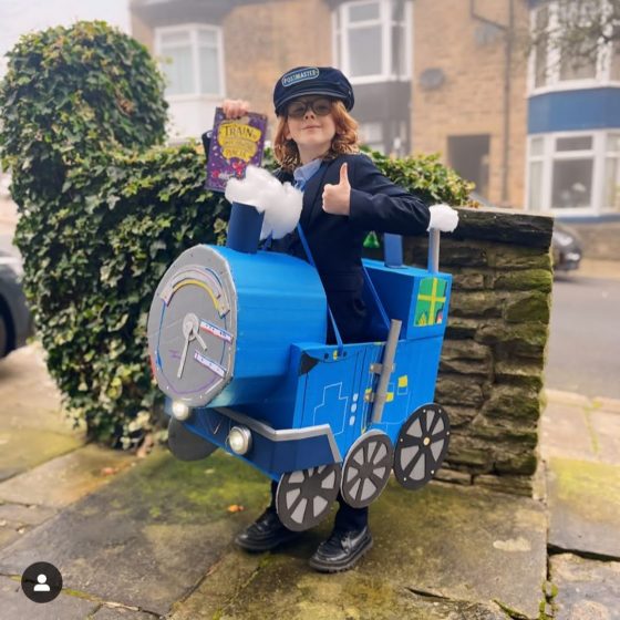 A young boy with ginger hair, wearing a navy blue postmaster's uniform and a large cardboard recreation of the Impossible Postal Express around his body. He holds a copy of The Train to Impossible Places and gives a thumbs up.