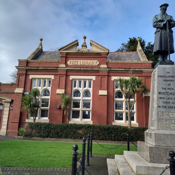 The exterior of Whitchurch Library. It is a small but handsome red brick building with palm trees, a box hedge and a small lawn outside. A war memorial with a bronze statue of a soldier stands in the foreground.