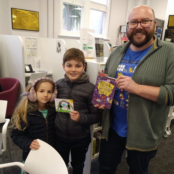 P.G. Bell poses with two young readers - a brother and sister in winter jackets. He holds a copy of The Train to Impossible Places. The young boy holds a drawing of two zombies he's made on a postcard.