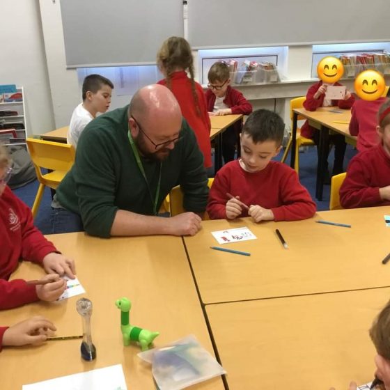 P.G. Bell sits with young school children in red jumpers. The children are drawing and colouring on blank postcards.