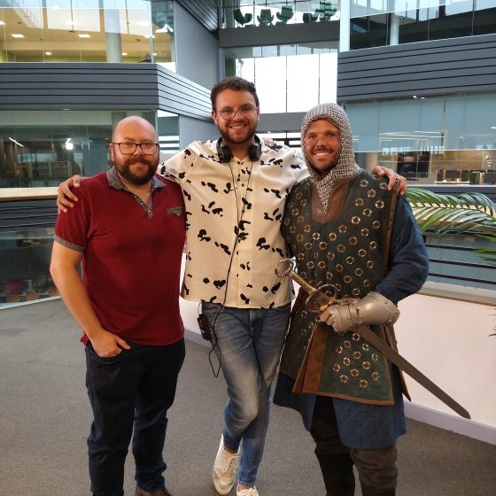 Three men stand on the balcony of the college library, overlooking the atrium. P.G. Bell is on the left- bald, bearded and wearing glasses, a burgundy polo shirt and jeans. Andrew Creak stands in the centre. He is taller than the other two, with glasses, a white shirt, jeans and headphones. Ben McGregor is on the right, dressed as a medieval knight with a sword.