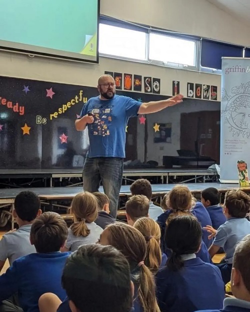 Author P.G. Bell, a rakishly good looking middle aged man with a beard and glasses, stands in front of a school hall full of primary children. The image catches him in the middle of a talk, unwittingly striking a dramatic pose with one arm extended towards the audience.